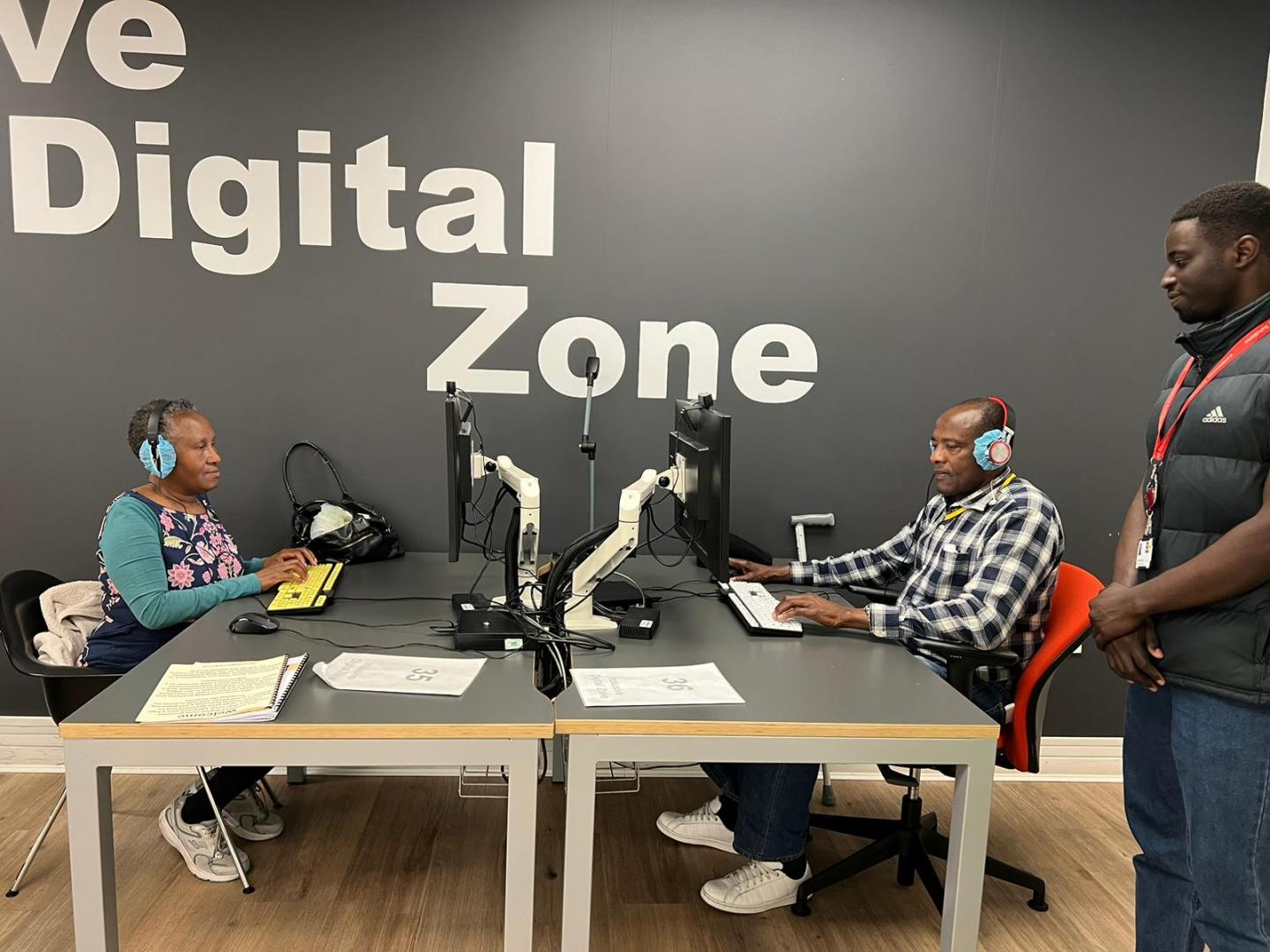 Two elderly people are sitting at a computer under an Inclusive Digital Zone sign and a man is standing next to them explaining how to use the computer