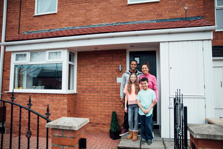 Photo of a family outside a house, it's a mum and dad and two children a boy and a girl and you can see the front door and front window and lower half of their house and a bit of their front garden