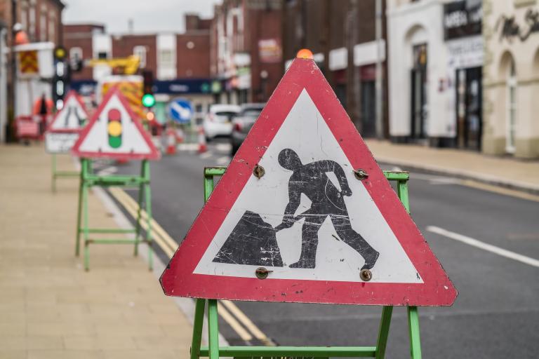 Photo of a roadworks sign and construction workers in the background on a road