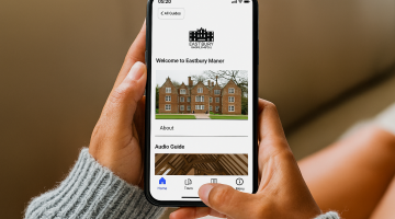 A photo of somebody holding a smartphone. Eastbury Manor House's website is on the screen with the text 'Welcome to Eastbury Manor' above an image of the house. Underneath the text says 'Audio guide'