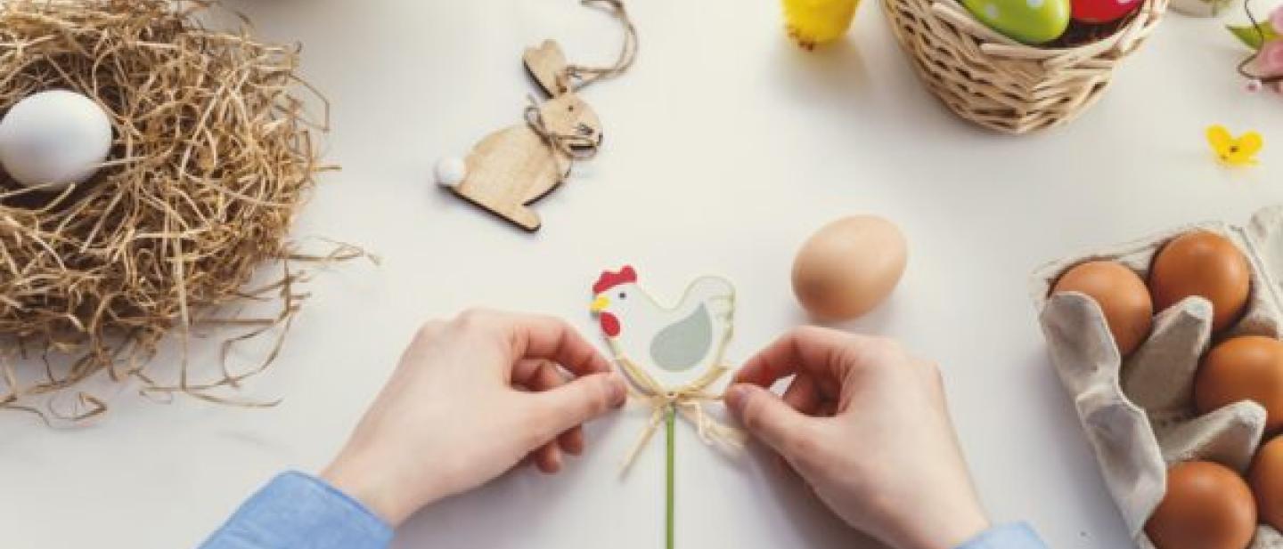 Two hands are tying a bow on an Easter chicken decoration. They are surrounded by items, on the right side there is a nest with a white egg, on the top there are two bowls with colourful eggs in them. On the left side there is a carton of brown eggs. Scattered on the table there are various small Easter decorations, such as a bunny and chicks.