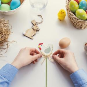 Two hands are tying a bow on an Easter chicken decoration. They are surrounded by items, on the right side there is a nest with a white egg, on the top there are two bowls with colourful eggs in them. On the left side there is a carton of brown eggs. Scattered on the table there are various small Easter decorations, such as a bunny and chicks.